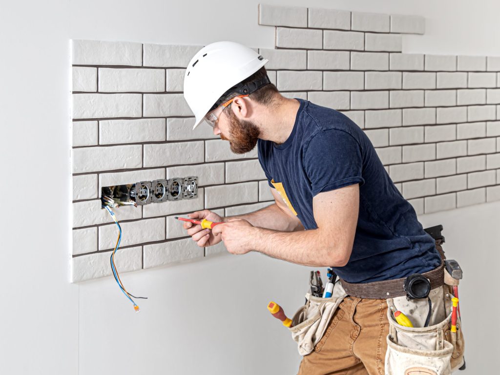 Electrician builder in a white helmet at work, installation of sockets, and switches. Electrician construction worker with a beard in overalls during the installation of sockets. Home renovation concept.